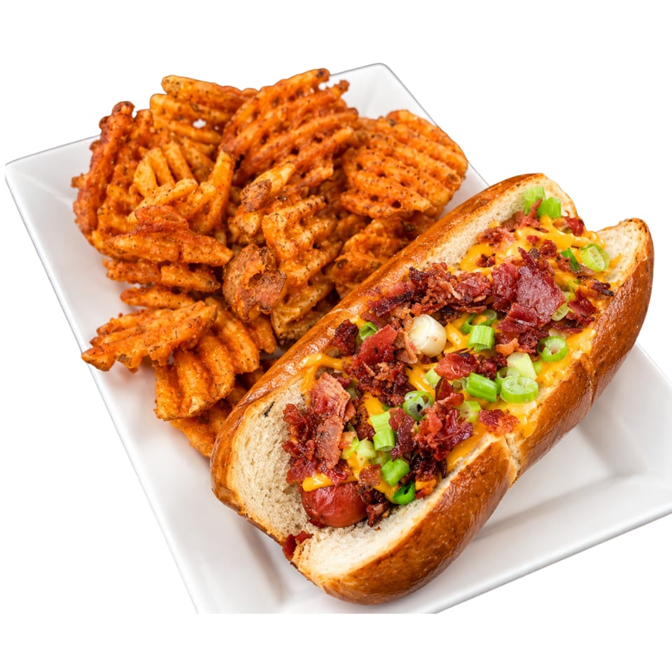 Loaded chili-cheese hot dog topped with bacon and green onions, served alongside seasoned waffle fries on a white plate.