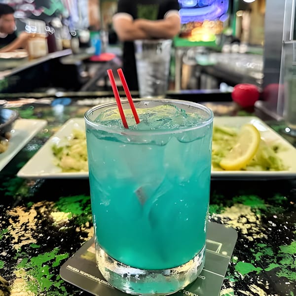 A vibrant blue cocktail with red straws sits on a bar, with food and a bartender in the background.